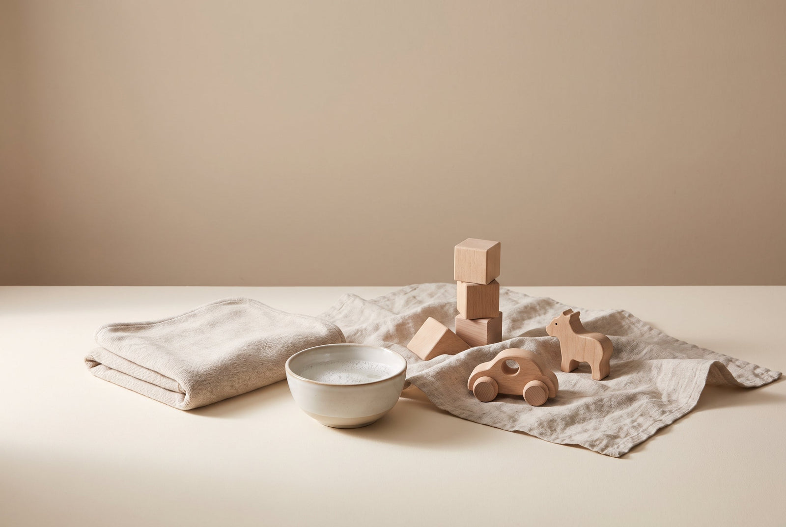 An adult cleaning a natural wooden toy with a soft cloth and a small bowl of mild soapy water on a cream surface