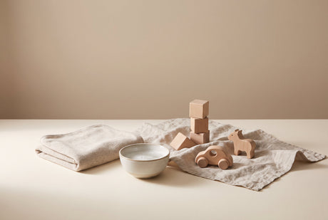 An adult cleaning a natural wooden toy with a soft cloth and a small bowl of mild soapy water on a cream surface