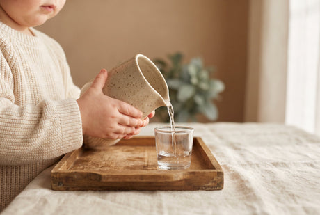 A toddler's hands pouring water from a small child-sized pitcher into a glass on a wooden tray, a practical-life activity
