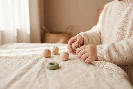 A toddler's hands carefully threading a wooden bead onto a lace on cream linen, showing calm concentration
