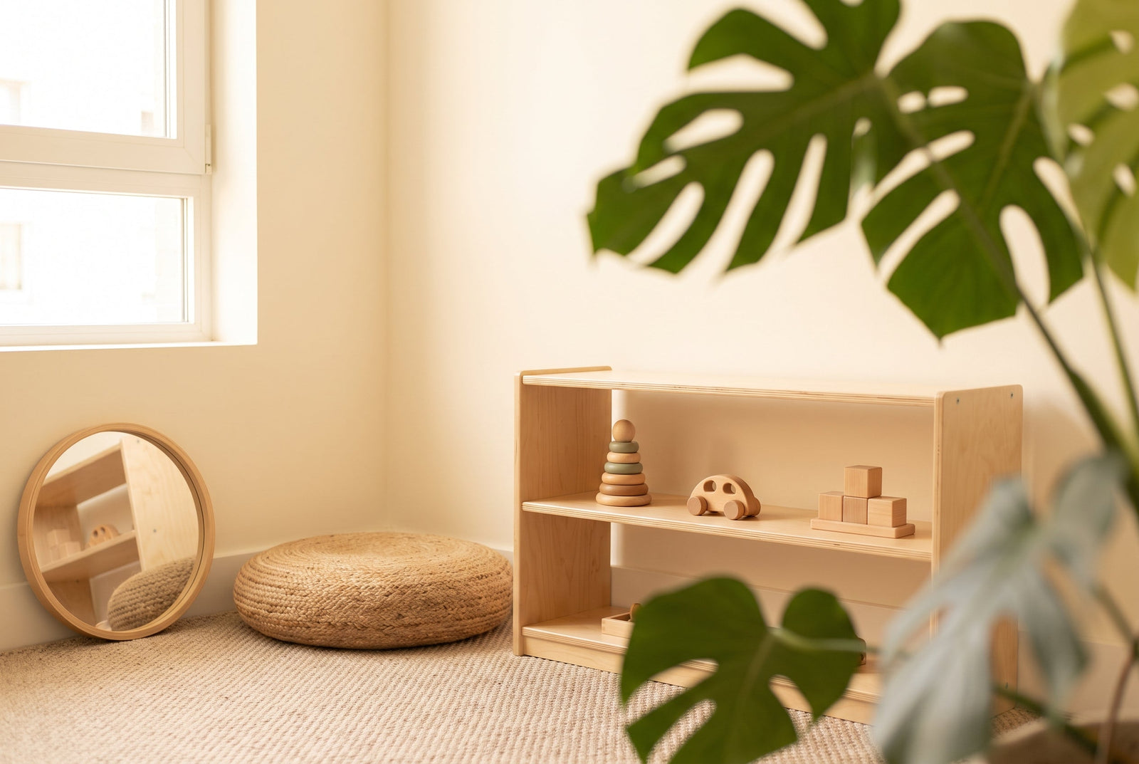 A low wooden Montessori shelf with a few natural toys, a floor cushion and a round mirror in a bright small-apartment corner