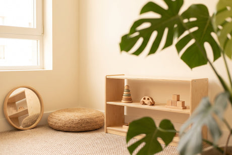 A low wooden Montessori shelf with a few natural toys, a floor cushion and a round mirror in a bright small-apartment corner