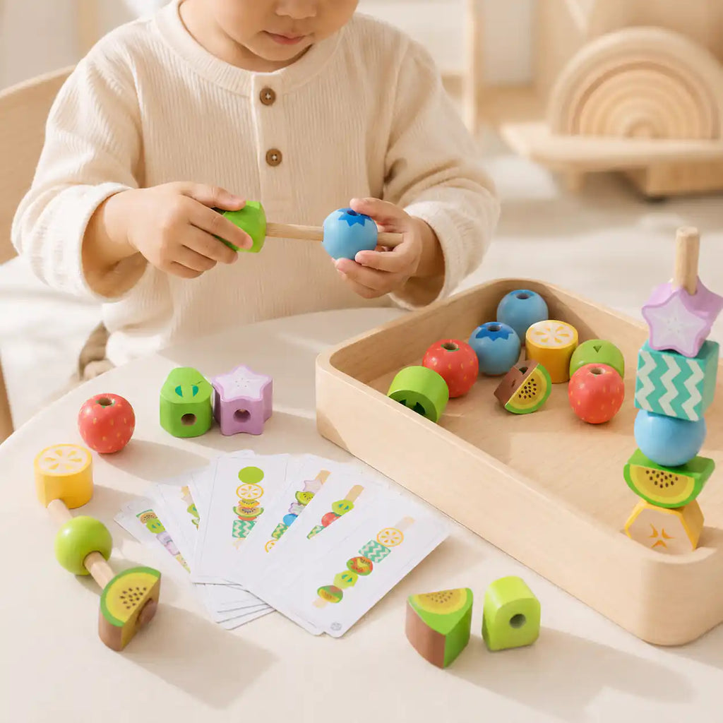Young child threading a wooden fruit bead onto a lacing stick, with a wooden tray of fruit beads and pattern cards on the table