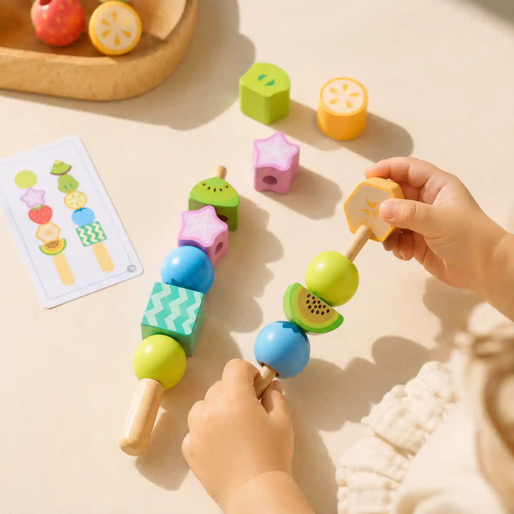 Toddler hands threading colorful wooden fruit beads onto two wooden lacing sticks, pattern card visible in the background