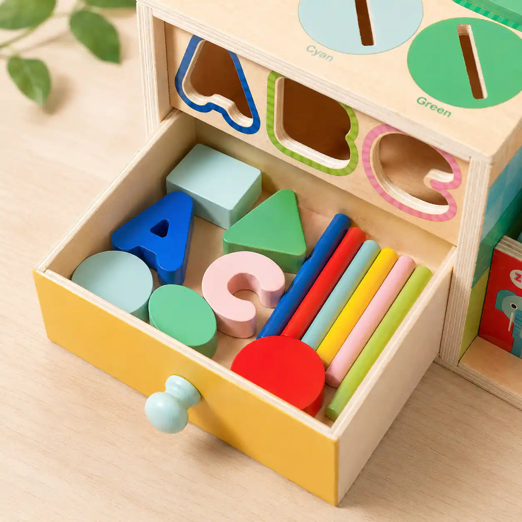 Top-down view of yellow storage drawer holding all wooden activity cube pieces including ABC letters, shapes, color coins, and rainbow sticks