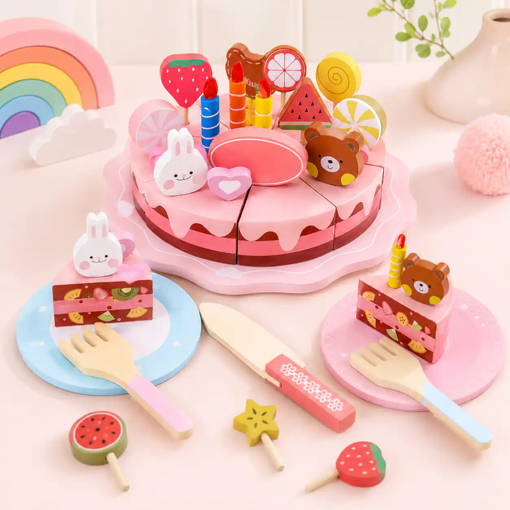 Wooden pretend play birthday cake styled with two decorated slices, fruit lollipops, and a wooden rainbow in the background