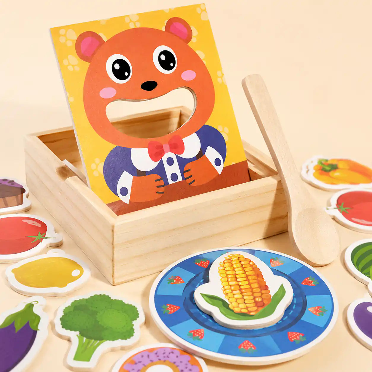 Wooden feed the bear toy with strawberry-patterned plate, corn piece, wooden spoon, and assorted wooden food pieces on a warm beige background