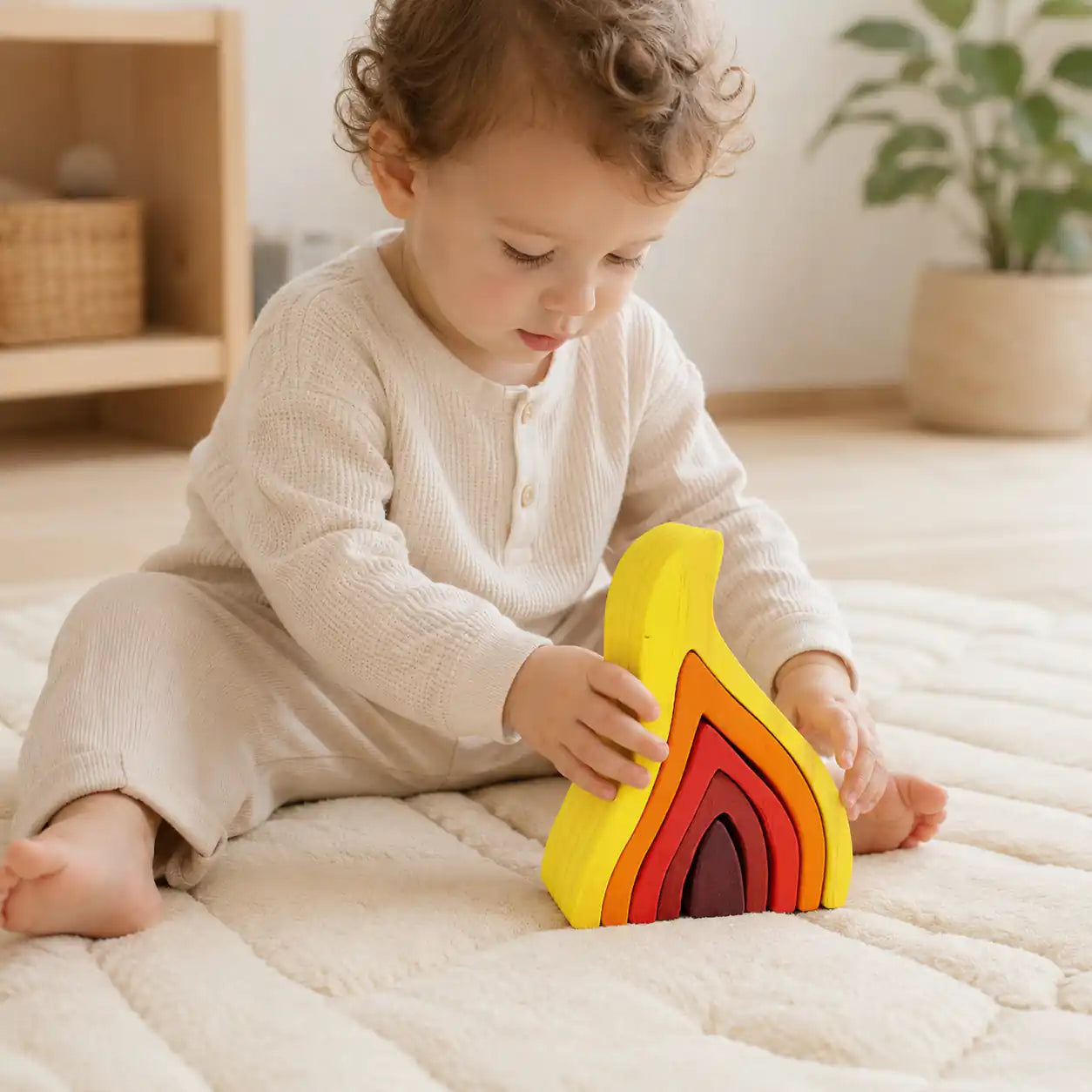 Curly-haired toddler in cream clothing sitting on a soft rug and playing with the wooden flame nesting puzzle