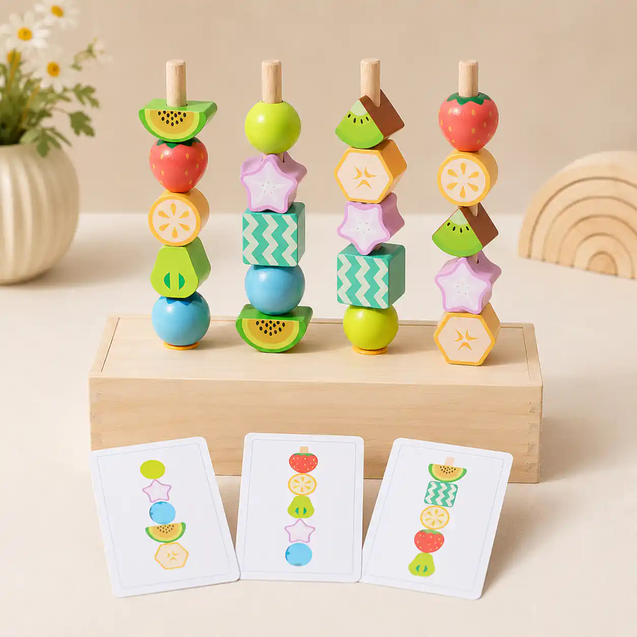 Four wooden dowels with stacked fruit beads on a wooden box base, with three pattern cards leaning in front showing bead sequences