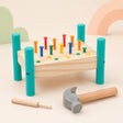 Wooden hammer and peg pounding bench with 14 colorful pegs upright, soft-headed hammer, and wooden punch tool on a cream nursery background with pastel cloud and rainbow shapes