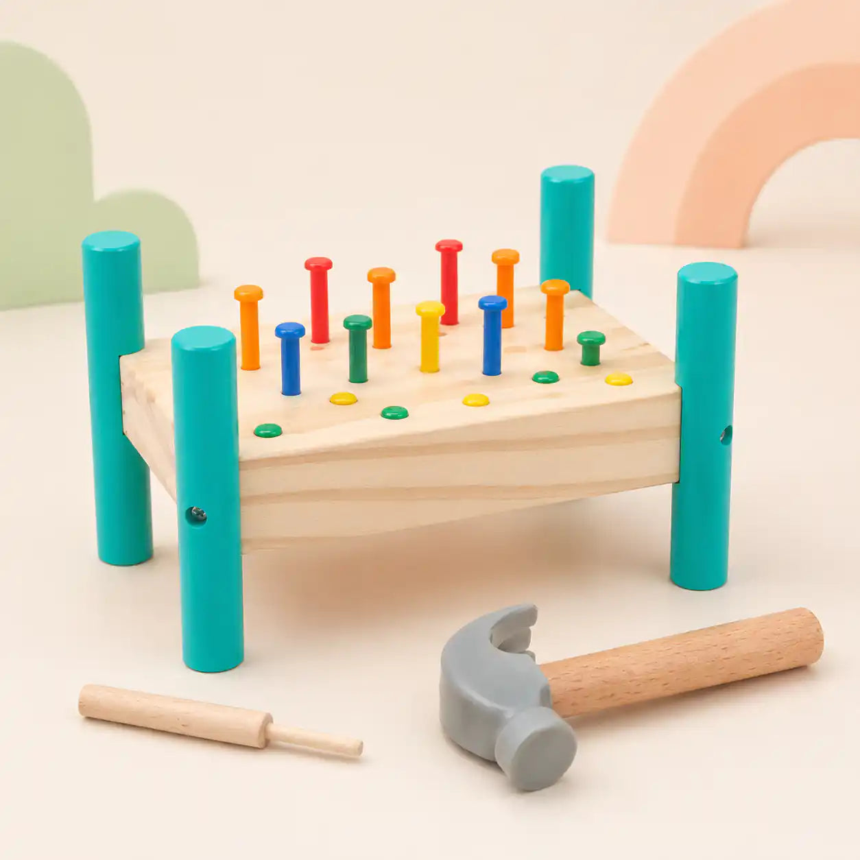 Wooden hammer and peg pounding bench with 14 colorful pegs upright, soft-headed hammer, and wooden punch tool on a cream nursery background with pastel cloud and rainbow shapes