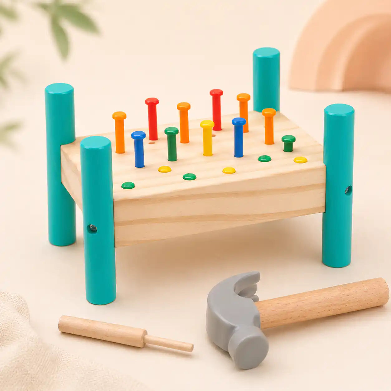 Side angle view of the wooden hammer and peg pounding bench showing teal painted legs, colorful pegs, hammer, and wooden punch tool on a warm cream background