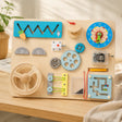 Wooden Montessori busy board with eleven hands-on activities including gears, clock and magnetic maze on a cream background