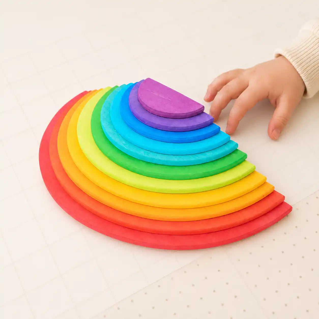 Toddler hand reaching for the smallest purple semicircle of a wooden rainbow nesting set on a soft mat