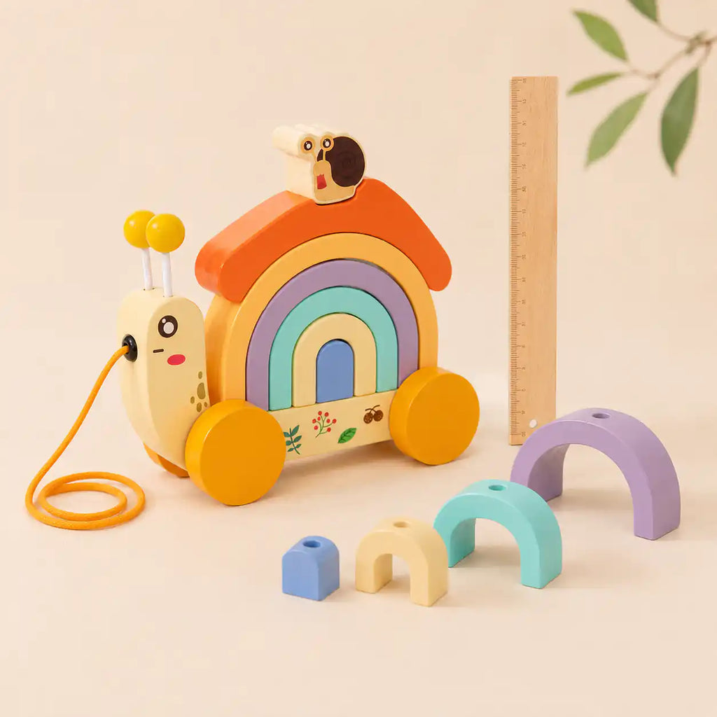 Wooden snail pull-along toy with pastel rainbow arches and small blue block laid out alongside on cream background